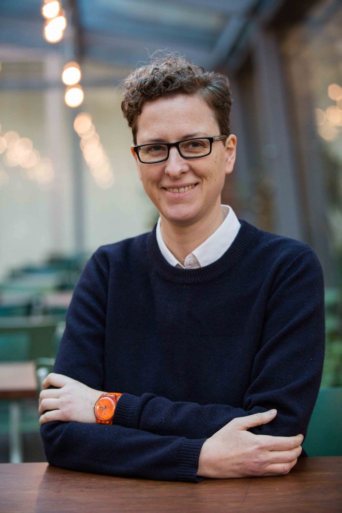The image shows Gitta, a white woman with short brown curly hair, dark-rimmed glasses. 
She is sitting at a desk with her arms folded in front of her. 
